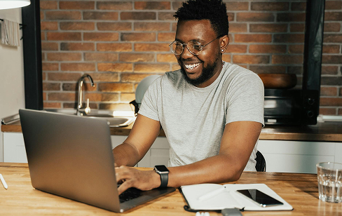 A smiling man in a gray shirt and glasses works on a laptop at a wooden kitchen table, with a smartphone, notebook, and glass of water nearby.