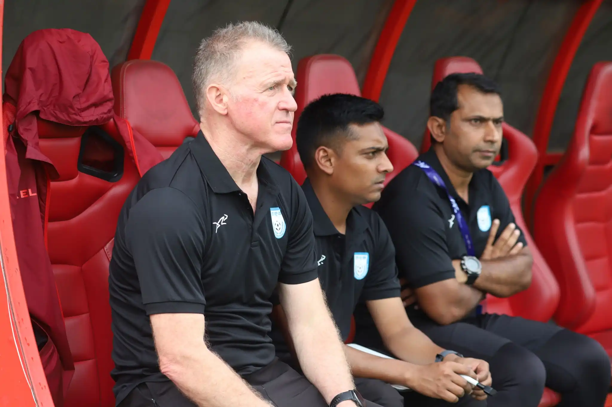 Bangladeshi women make history by qualified for the AFC Asian Cup football 3 Three coaches in black shirts sit on a red bench, focusing intently. Their expressions show concentration and anticipation during a sports event.